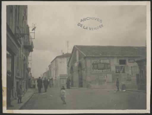 Une rue, avec des passants, des commerces et de nombreuses publicités collées sur le mur des halles (dont une grande pour le petit-beurre Lefèvre-Utile).